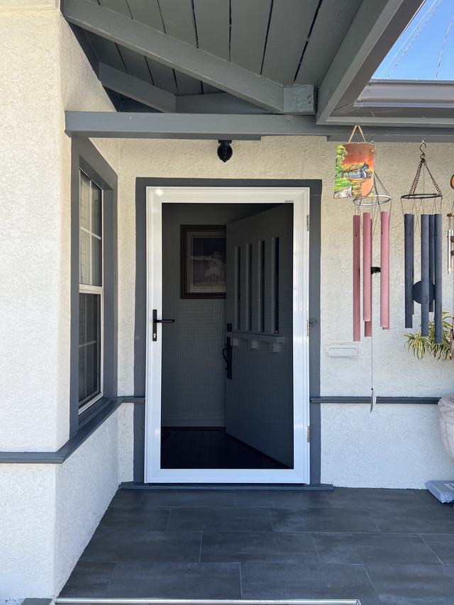 White-framed hinged security screen door on a covered stucco porch, with the front door open behind it