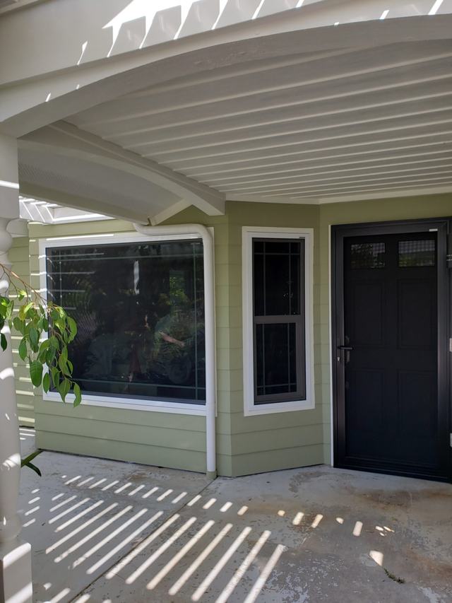 Black hinged security screen door beside screened windows on a green home, under a white patio cover